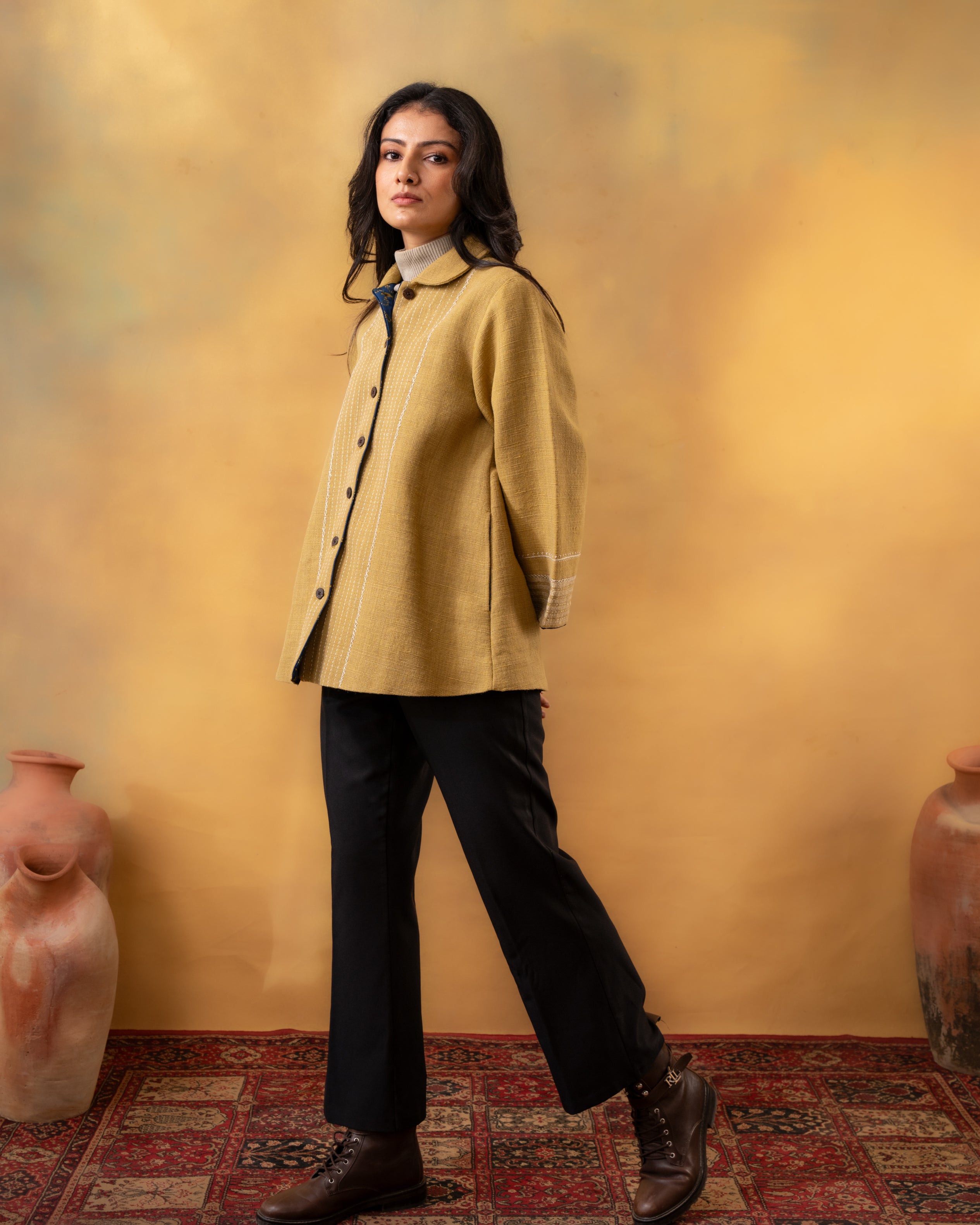 Woman wearing a mustard yellow coat against a warm-toned wall with decorative vases.